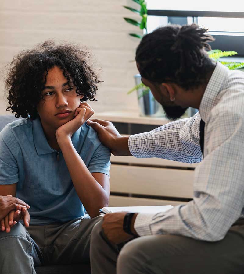 A young kid with his hand holding his head looks at a man who has his hand on the boy's shoulder.