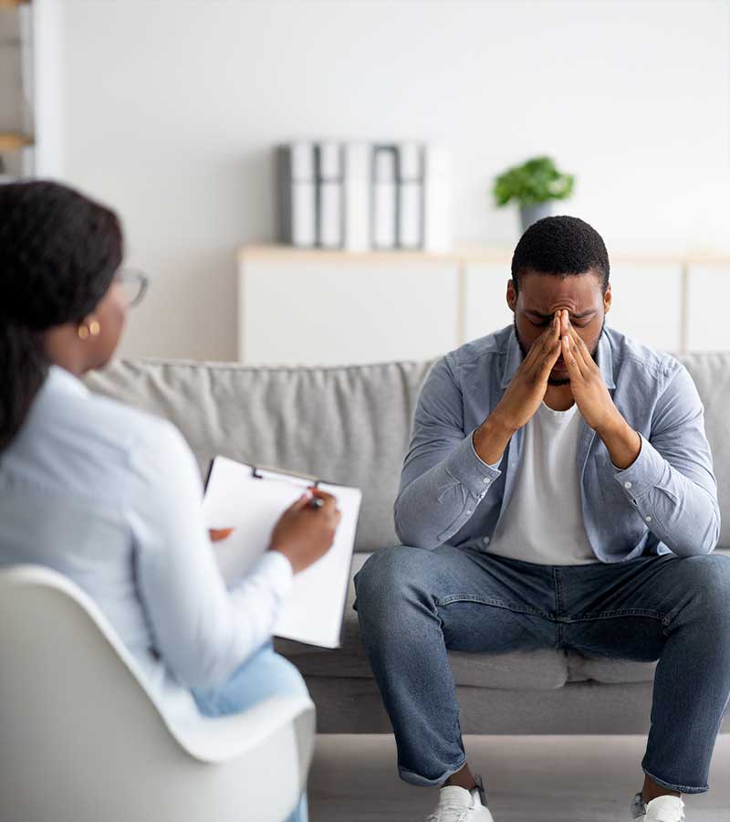 A young man sits on a sofa with his fingers tented and his head resting on them as he listens to a woman with a chart speaking to him.