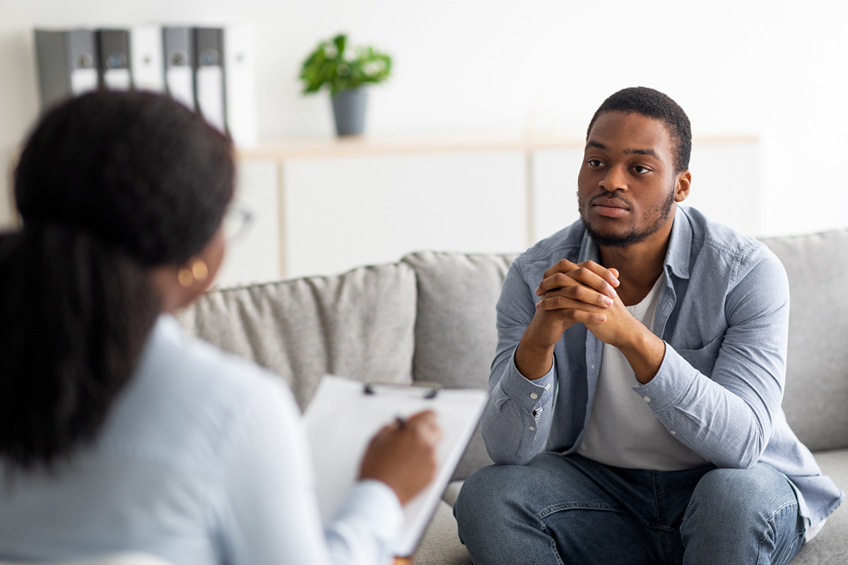 A man sitting with his hands intertwined looking with interest at a person with a clipboard talking.